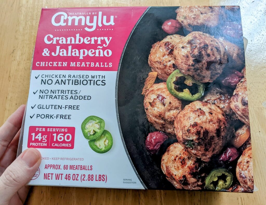 A hand holding a box of Amylu Cranberry & Jalapeno Chicken Meatballs from Costco.