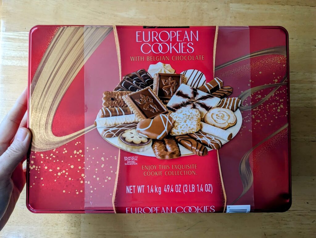 A hand holding a red rectangular metal tin of Costco Kirkland European Cookies on a wooden table.