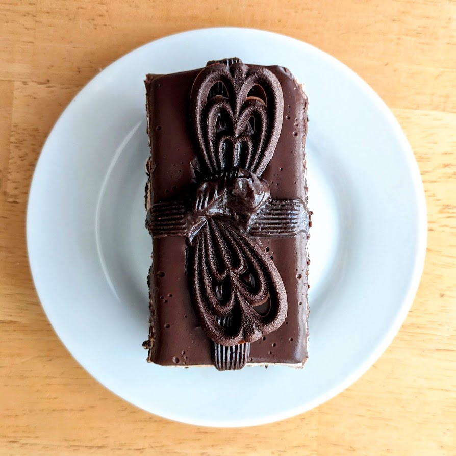 A slice of chocolate ganache supreme cake from the Publix Bakery, sitting on a white plate on a wood table.