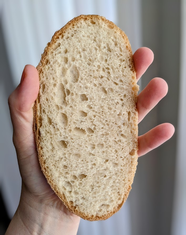 A slice of Costco bakery sourdough bread in a hand, held vertically in front of a gray background.