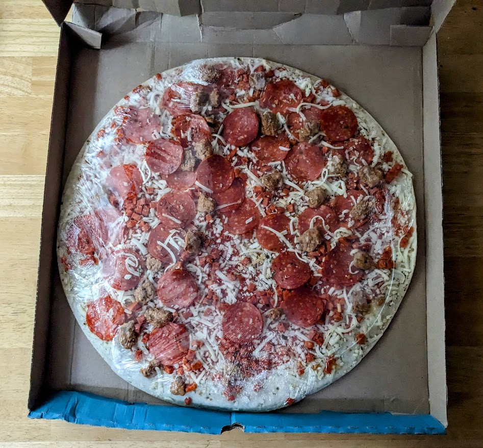 An open cardboard pizza box with a shrink wrapped sausage and pepperoni thin crust take & bake from Aldi Mama Cozzi's inside.