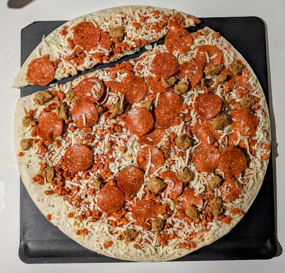 An extra large sausage and pepperoni take and bake thin crust pizza from Aldi, sitting on a gray baking sheet, with a corner of the crust broken off.