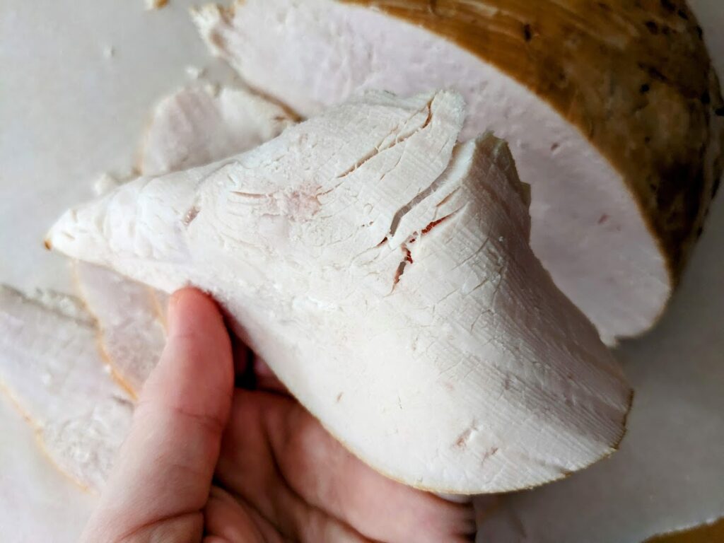 A hand holding a slice of Costco Kirkland Oven Browned Turkey Breast sliced thinly.