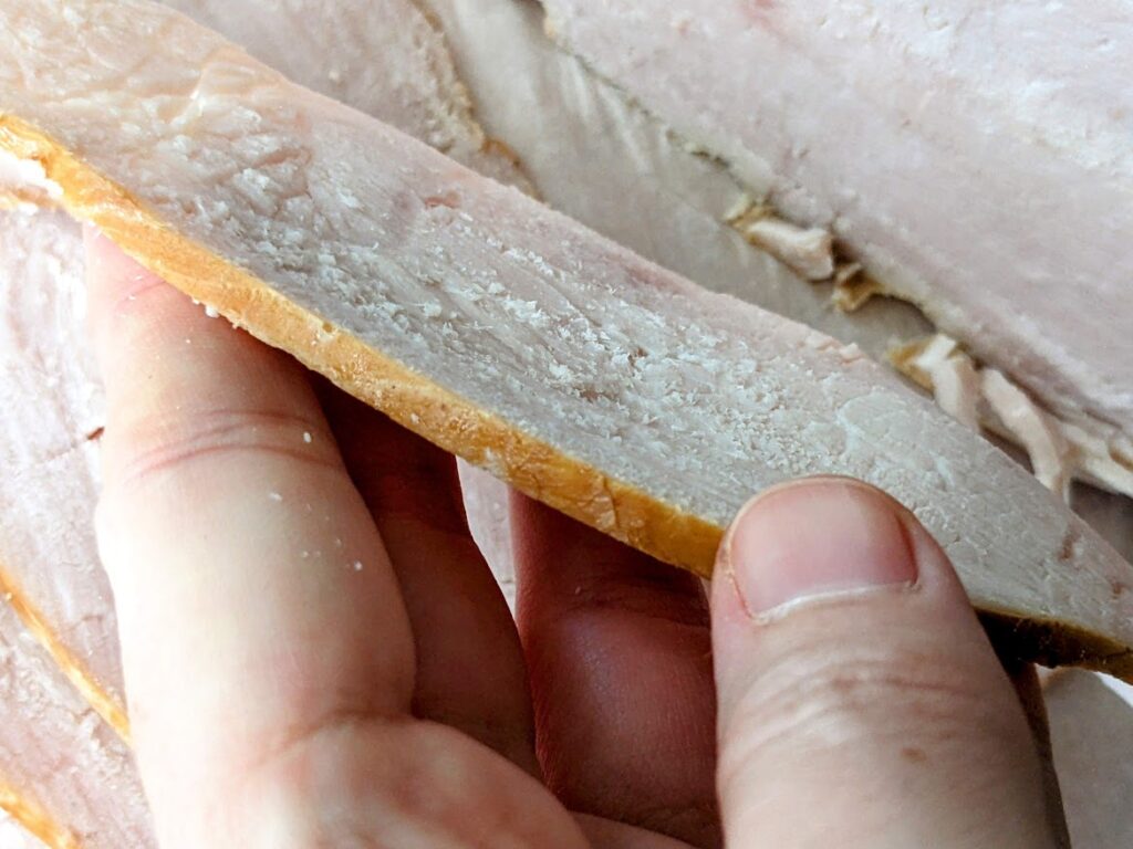 A hand holding a thin-sliced piece of Kirkland Oven Browned Turkey Breast from Costco.