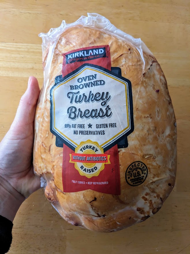 A hand holding a shrink-wrapped whole turkey breast from Costco Kirkland brand over a wood table.