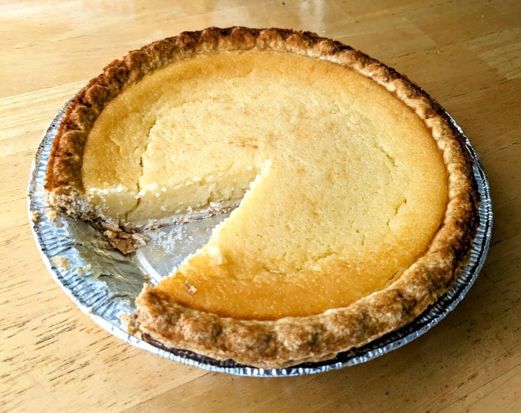 A whole Buttermilk Pie from Publix, with a slice removed, sitting in an aluminum pie pan on a wood table.