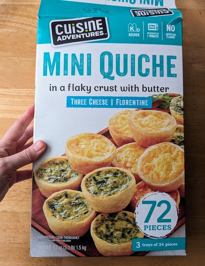 A hand holding a box of frozen mini quiches from Cuisine Adventures, purchased at Costco.