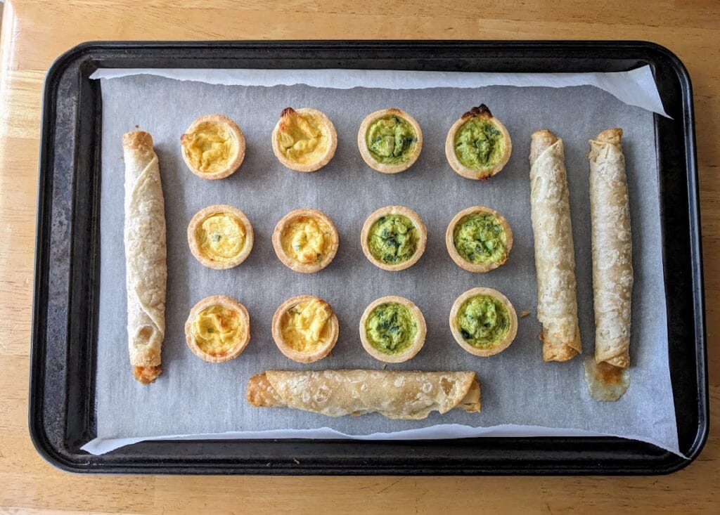 A baking pan with parchment paper and rows of mini quiches and taquitos appetizers.