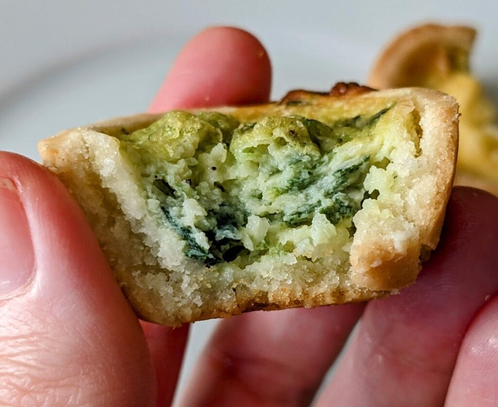 A hand holding a bitten Cuisine Adventures Florentine spinach mini quiche.