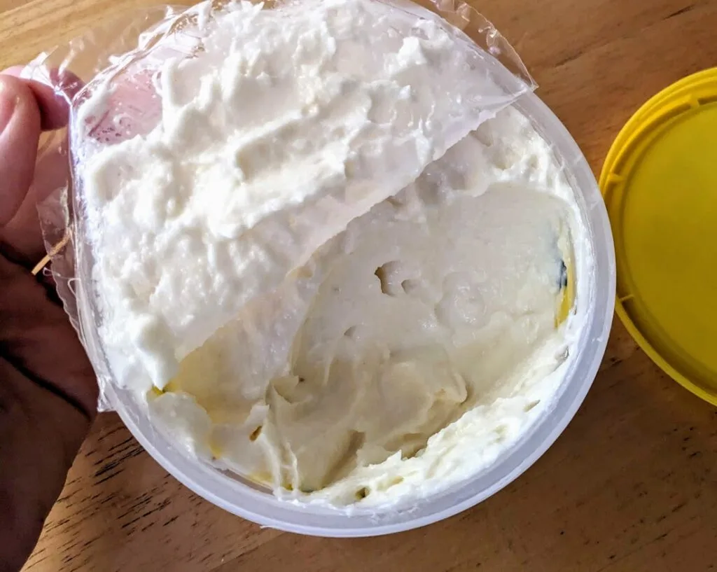 A hand peeling back the plastic film over a tub of TOOM garlic dip.