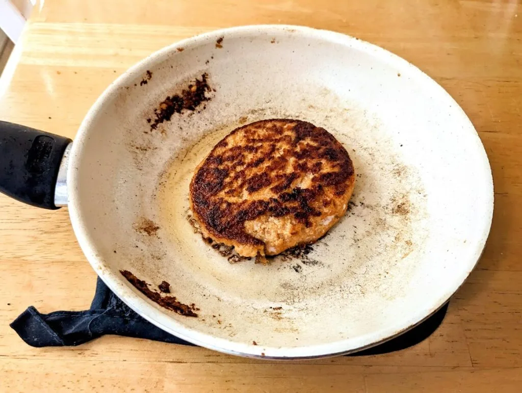 A ceramic frying pan with a browned salmon burger patty from Aldi, sitting on a kitchen table.