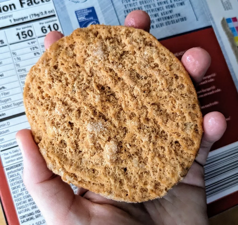 A hand holding a frozen Fremont Chipotle Alaska Salmon Burger patty from Aldi.