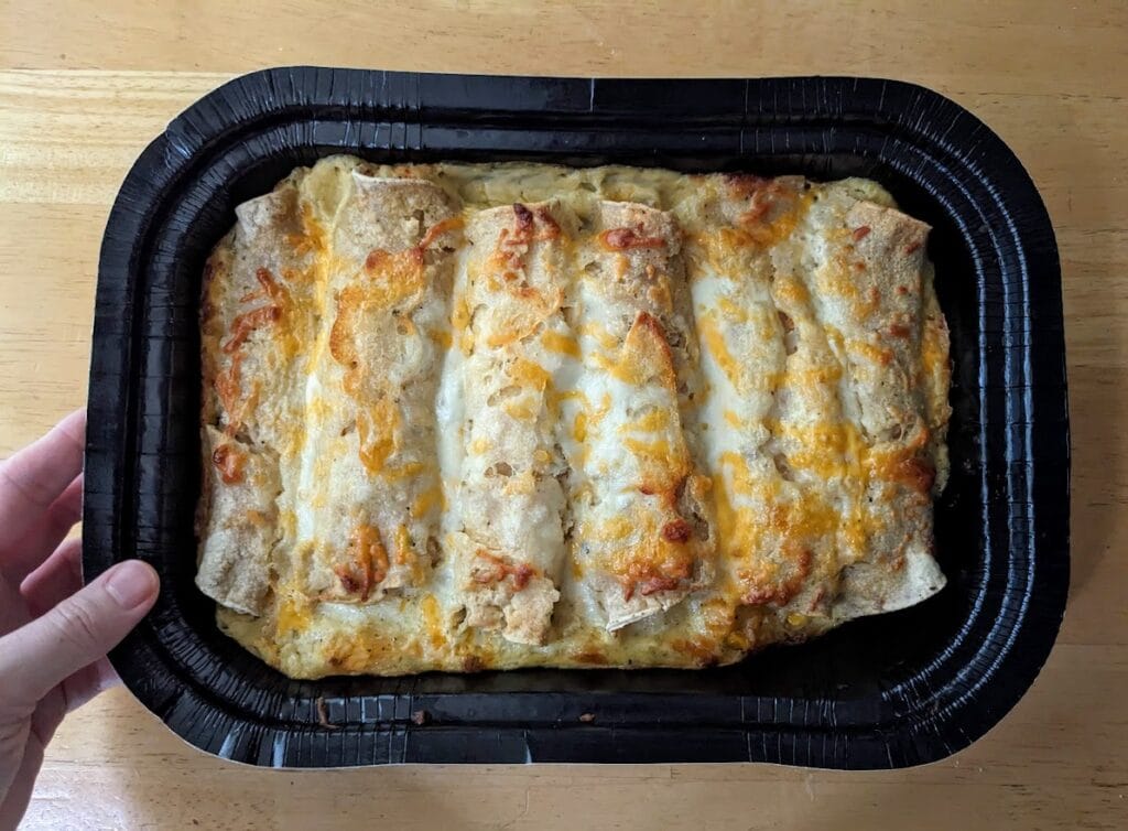 A hand holding a black cardboard tray of baked chicken and cheese enchiladas from Chef Gustavo, purchased from Costco.