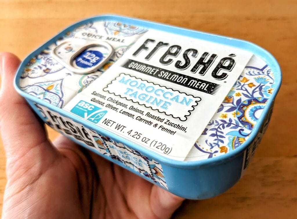 A hand holding a blue can of Freshe Gourmet Salmon Meal Moroccan Tagine.