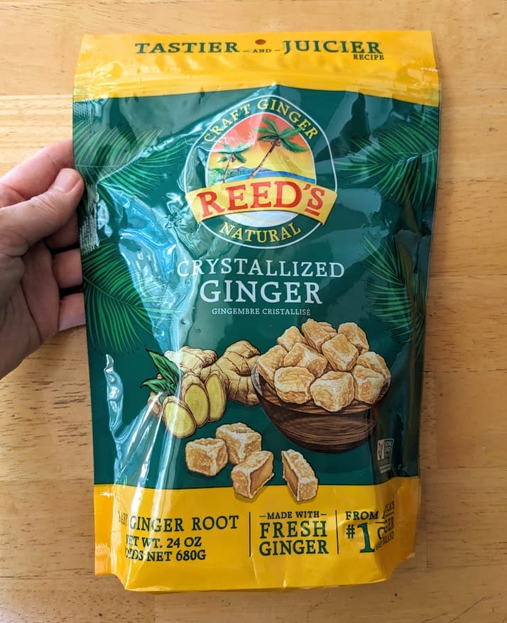 A hand holding a plastic bag of Reed's Natural Crystalized Ginger from Costco over a kitchen table.