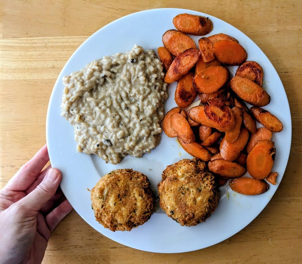 A hand holding a white plate with cooked carrots, crab cakes, and Priano Mushroom Risotto from Aldi.