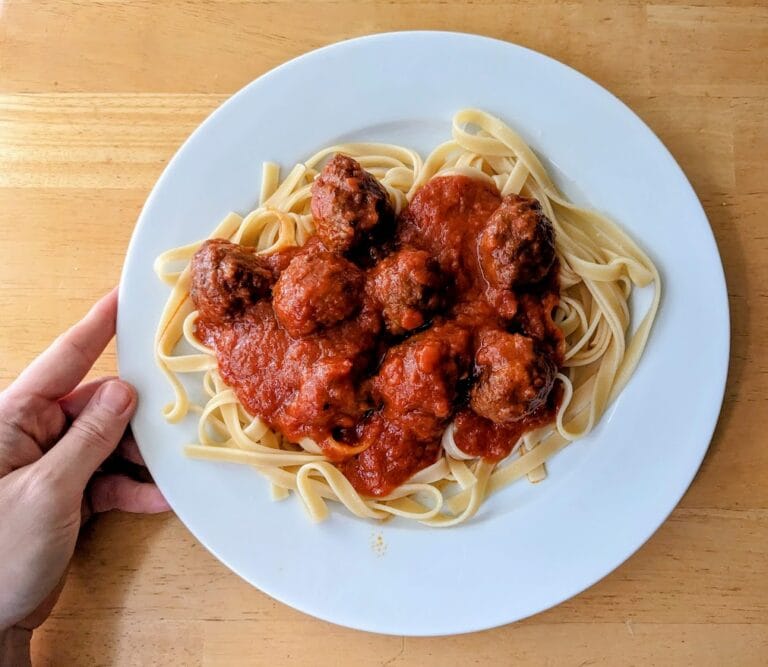 A hand holding a white plate with pasta topped with sauce and Kirkland Italian Style Beef Meatballs from Costco.