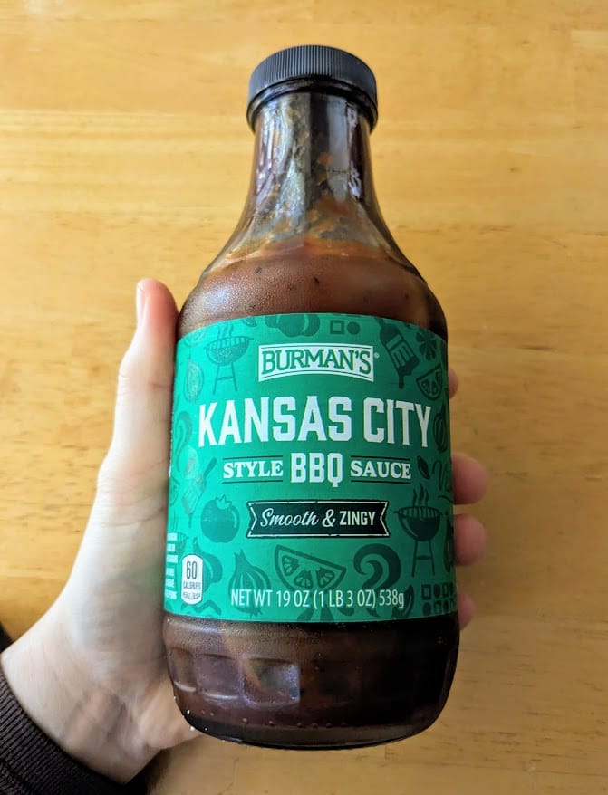 A hand holding a glass bottle of Burman's Kansas City Style BBQ Sauce from Aldi.
