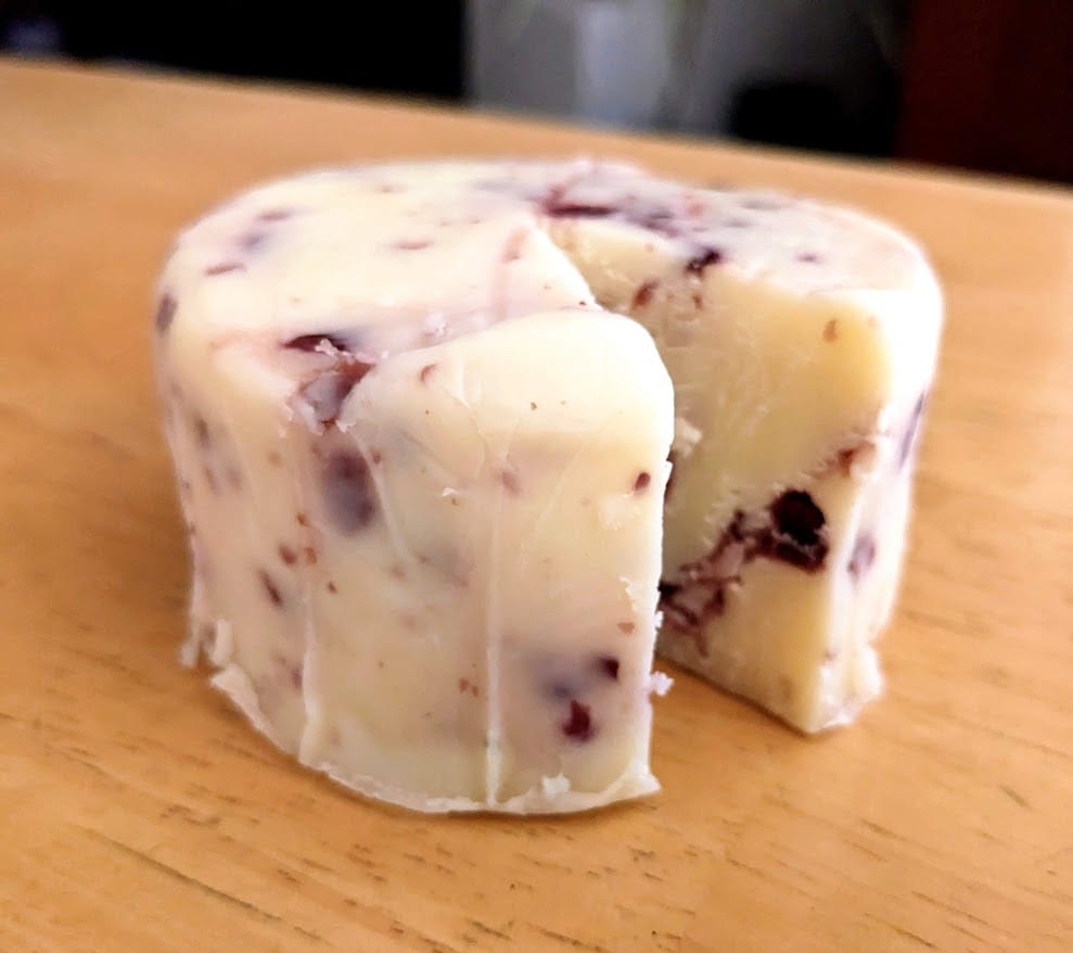 A circular block of Aldi Cranberry White Cheddar Cheese sitting on a kitchen table.