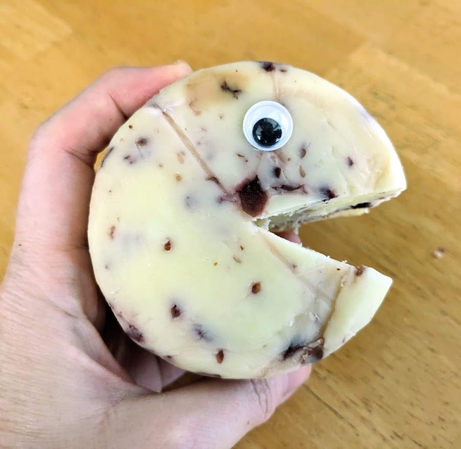 A hand holding a circle of Aldi Cranberry White Cheddar cheese with a googly eye and a wedge removed to look like a mouth.