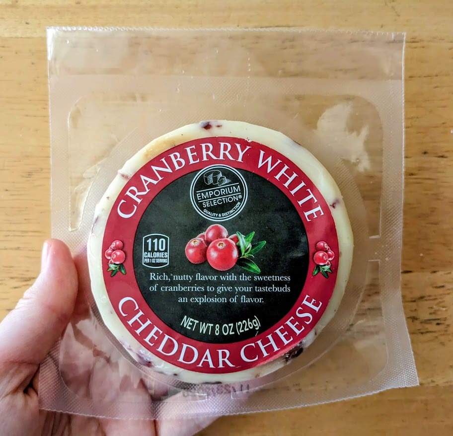 A hand holding a package of Cranberry White Cheddar Cheese from Aldi Emporium Selected store brand.