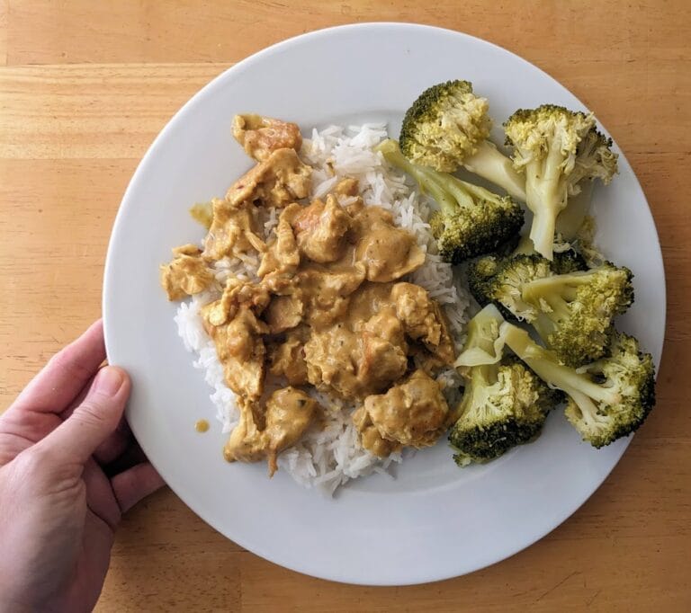A hand holding a white plate with rice, broccoli, and Kevin's Thai Coconut Curry Chicken meal.