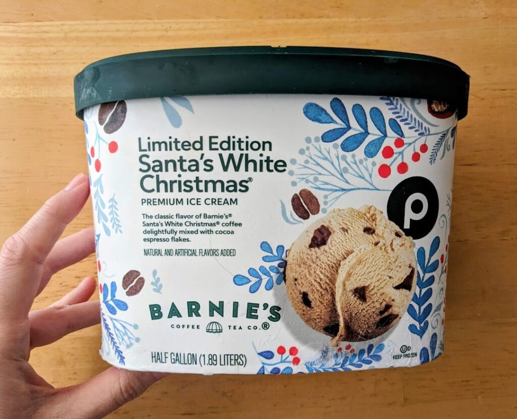 A hand hold a half gallon tub of Publix Limited Edition Santa's White Christmas Premium Ice Cream with Barnie's Coffee.