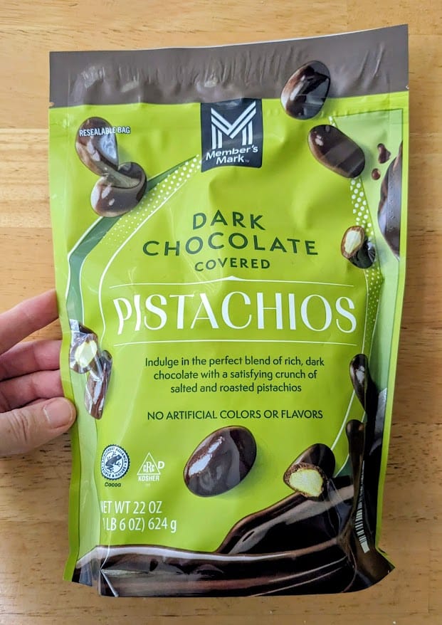 A hand holding a bag of Dark Chocolate Covered Pistachios from Sam's Club.