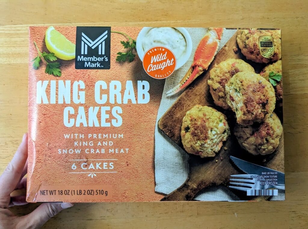 A hand holding a Member's Mark frozen King Crab Cakes package.