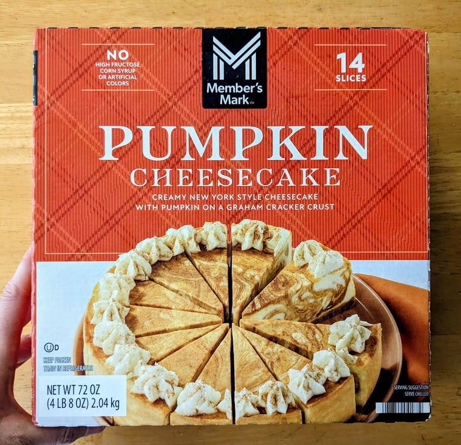 A hand holding a box of frozen Pumpkin Cheesecake from Sam's Club Member's Mark store brand.