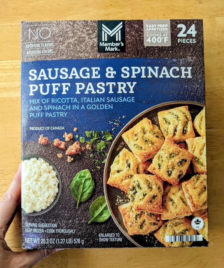 A hand holding a box of frozen Sausage and Spinach Puff Pastry appetizers from Sam's Club.