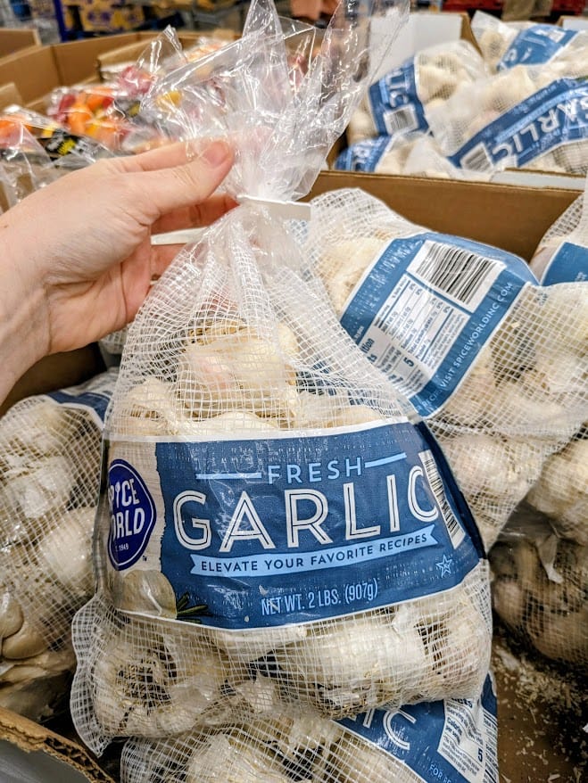 A hand holding a bag of whole garlic bulbs inside a Sam's Club warehouse.