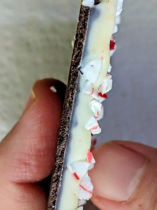 Fingers holding the edge of a piece of white and dark chocolate layered Peppermint Bark from Costco.