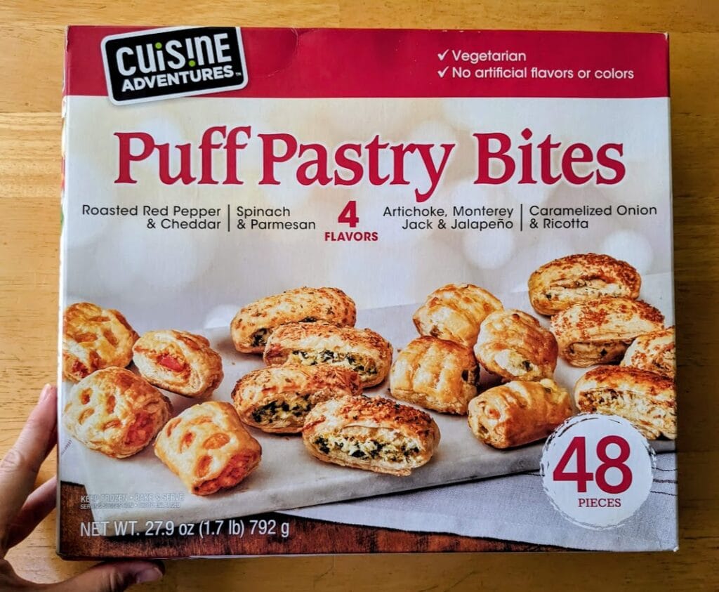 A hand holding a box of frozen Cuisine Adventures Puff Pastry Bites appetizers from Costco.