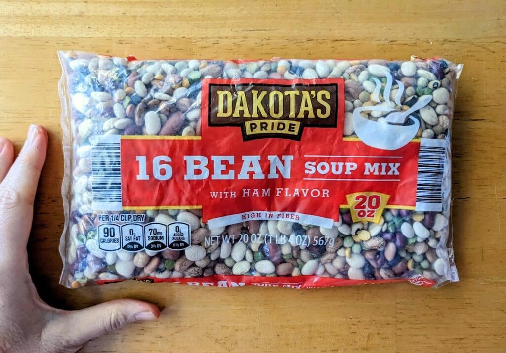A hand next to a 20 ounce bag of Aldi Dakota's Pride 16 Bean Soup Mix with Ham Flavor.