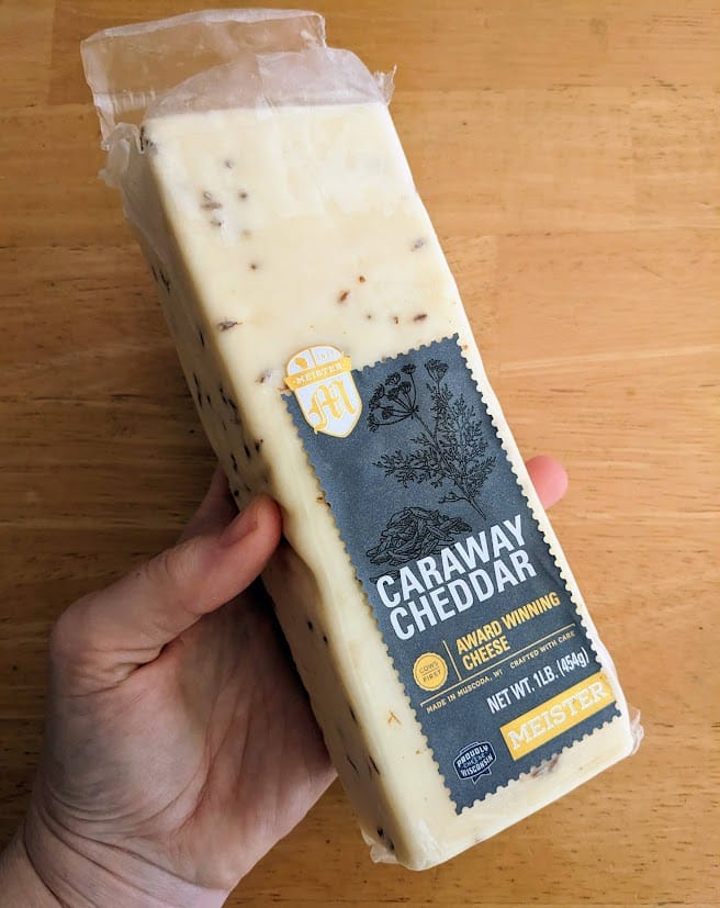 A hand holding a block of Meister Caraway Cheddar cheese over a kitchen table.