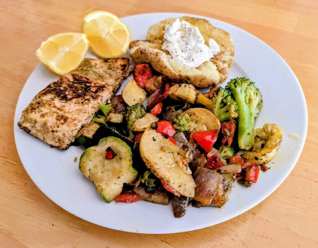 A white plate with a serving of salmon, baked potato, lemon slices, and cooked Primavera Mistura from Pura Vida.