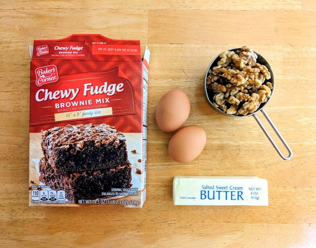A box of Aldi Baker's Corner Chewy Fudge Brownie Mix on a kitchen table with two eggs, a stick of butter, and a measuring cup of walnuts.