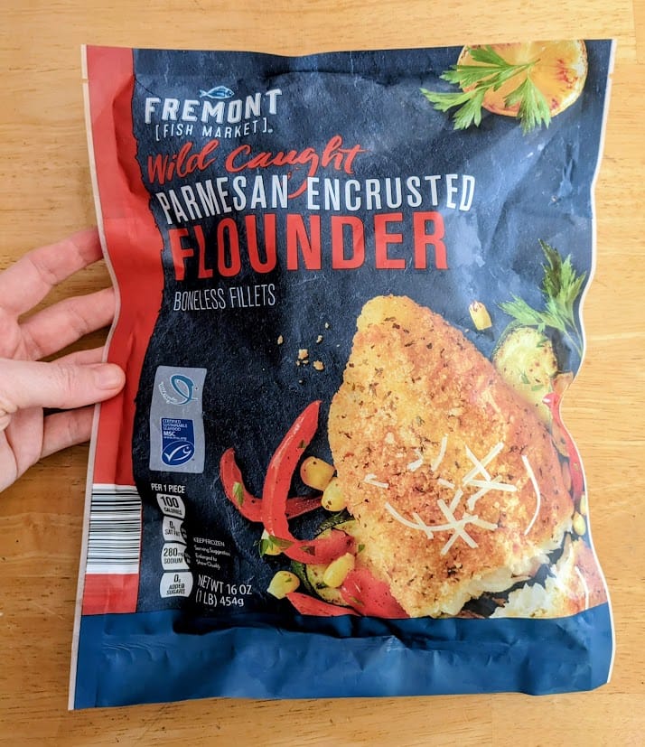 A hand holding a bag of frozen Fremont Fish Market Wild Caught Parmesan Encrusted Flounder Boneless Fillets from Aldi.