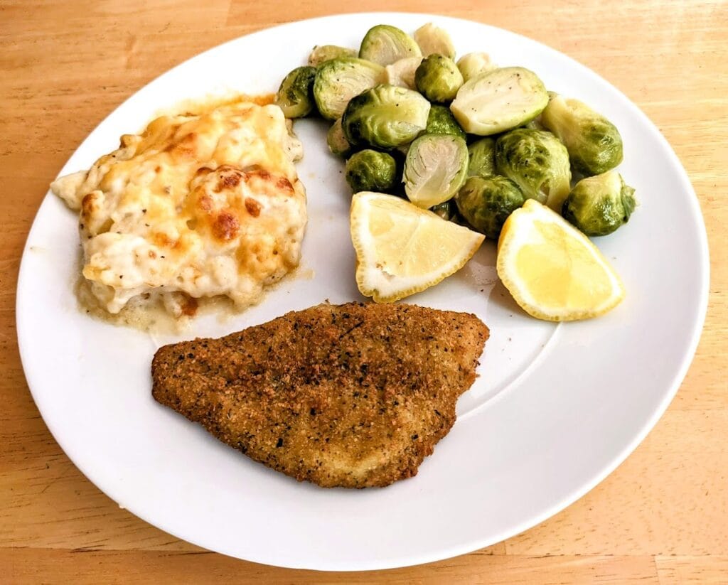 A white plate with a serving of Aldi Parmesan Crusted Flounder, brussels sprouts, lemon wedges, and scalloped potatoes.