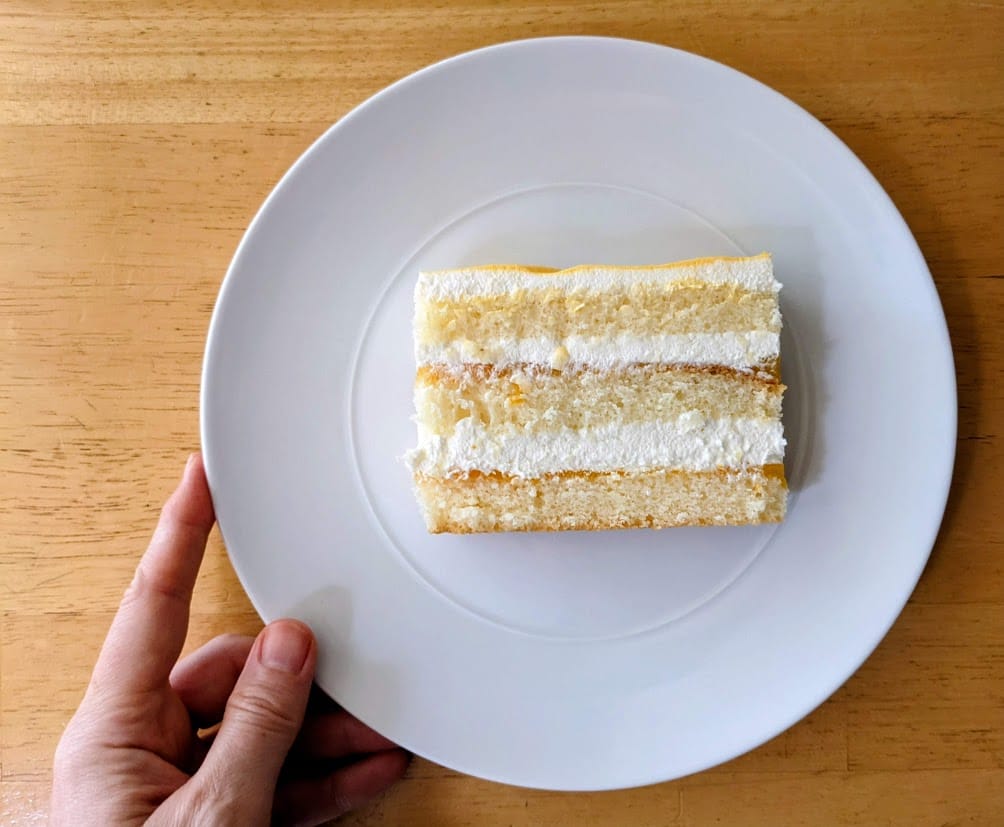 A hand holding a white plate with a slice of Costco Peaches and Cream Bar Cake.