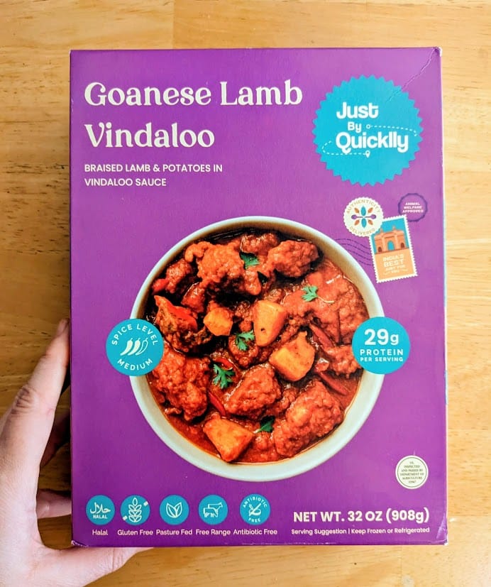 A hand holding a box of Goanese Lamb Vindaloo curry from Quicklly Indian Food.