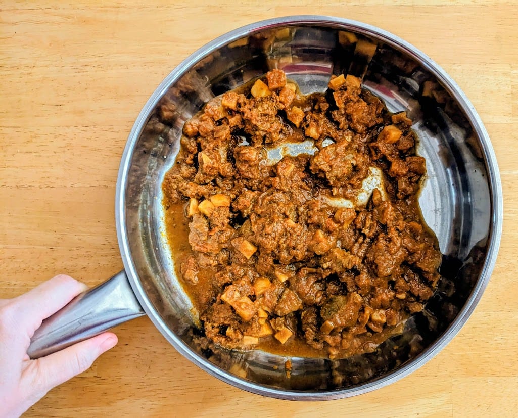 A hand holding a stainless steel pan with Quicklly Indian Lamb Vindaloo curry.