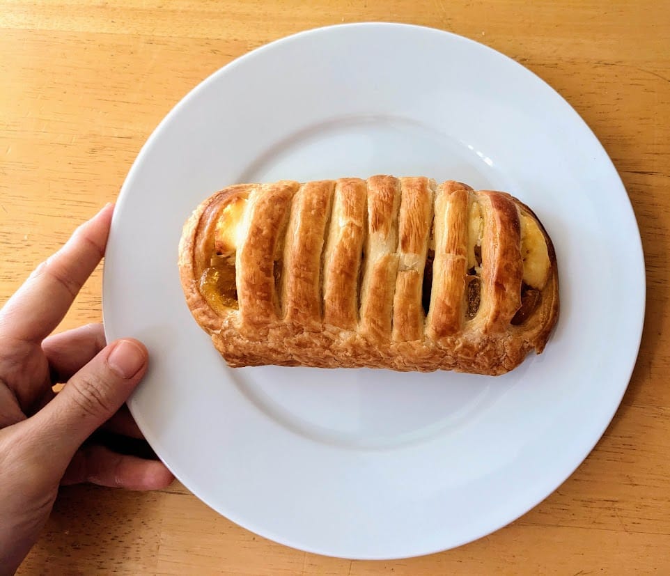 A hand holding a white plate with a whole Costco Peaches and Cream Pastry.