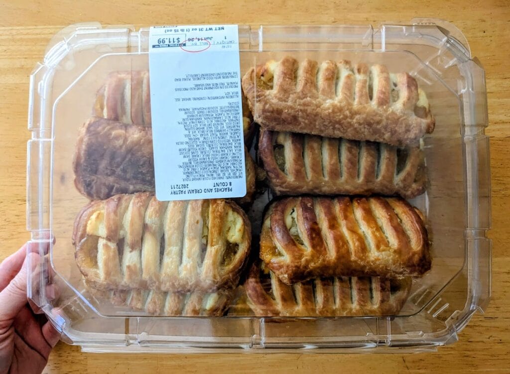 A hand holding a plastic box of Peaches & Cream pastries from the Costco Bakery.