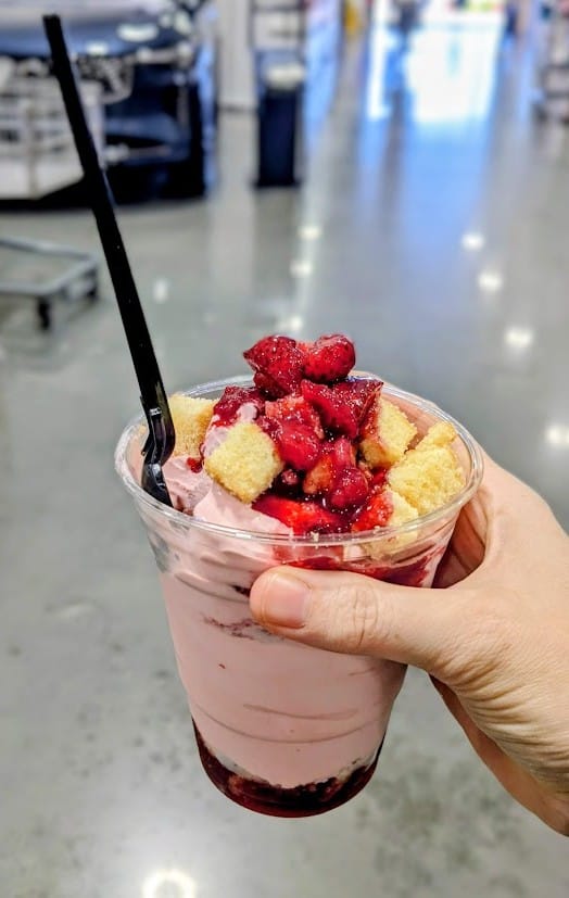 A hand holding a plastic cup of Strawberry Shortcake Sundae inside a Costco warehouse.