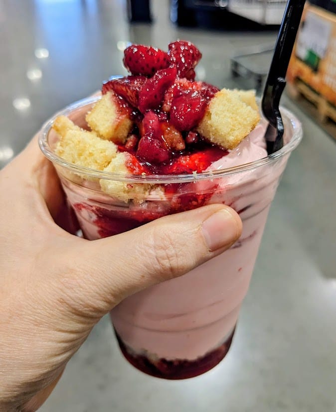 A hand holding a plastic cup of pink Strawberry Shortcake Sundae topped with fruit from the Costco Food Court.