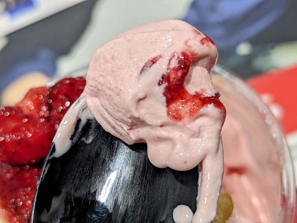 A plastic spoon with a bite of pink ice cream and fruit sauce from a Costco Strawberry Shortcake Sundae.