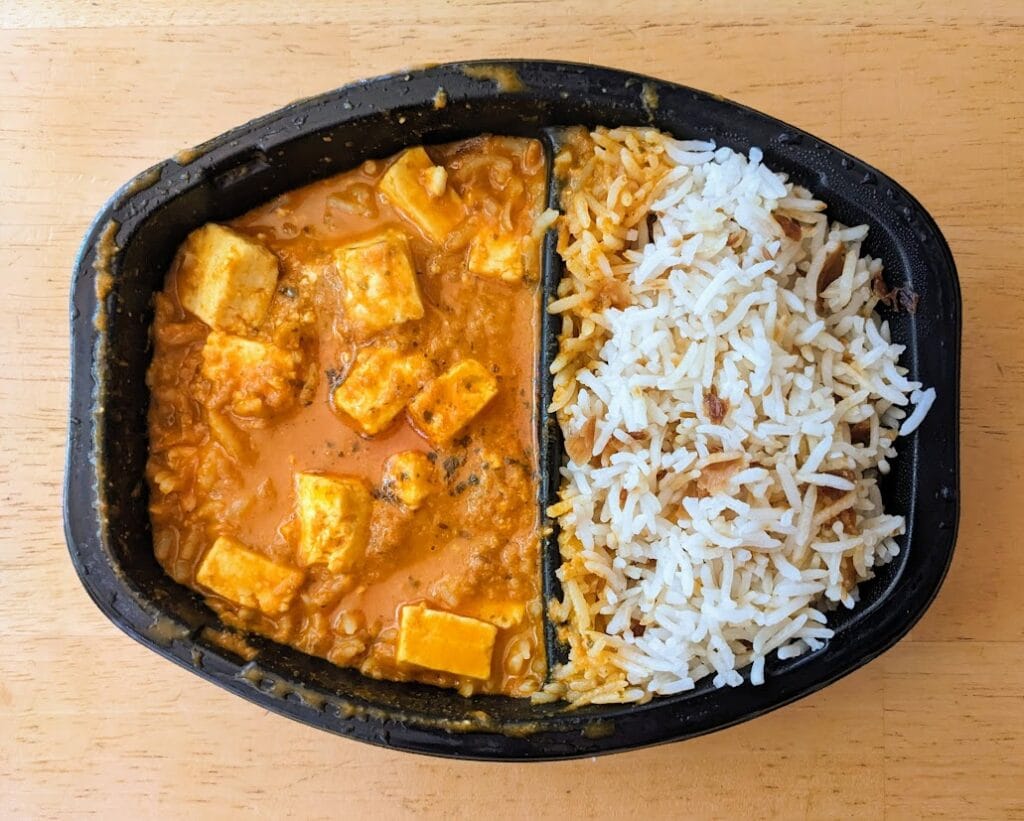 A microwaved black plastic tray of Paneer Tikka Masala and Onion Basmati Rice from Deep Indian Kitchen.