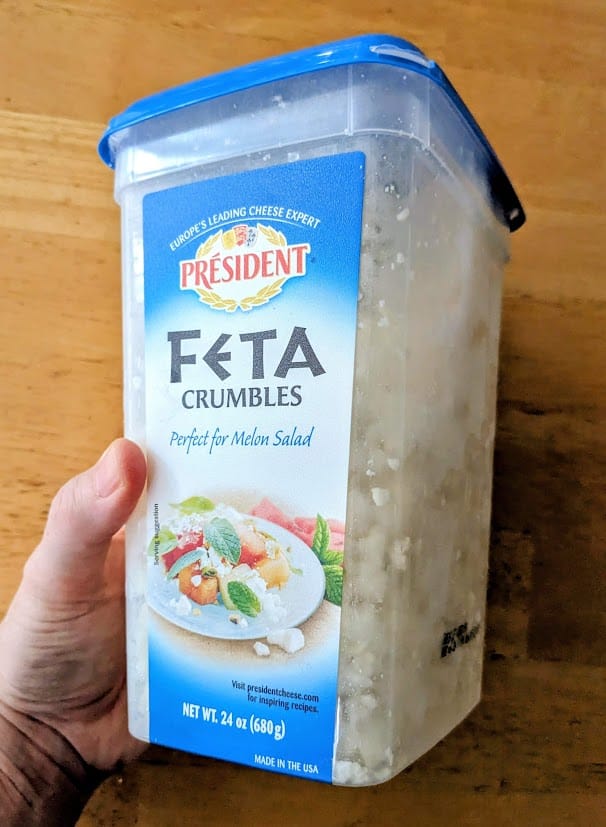 A hand holding a plastic tub of President brand Feta cheese Crumbles from Costco.